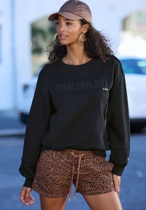Schwarzer Sweatshirt mit dem Text "DREAM wild", kombiniert mit braunen Leopardenprint-Shorts. Enthält goldene Akzente und eine braune Kappe. Glatte Stofftextur.