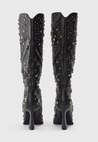 Botas de rodilla de tacón alto negras con tachuelas de metal plateado y patrones decorativos, vistas desde la parte de atrás sobre un fondo blanco.