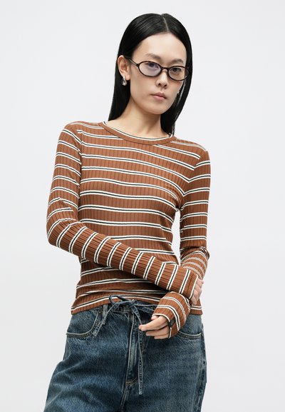 Camisola de manga comprida às riscas em tecido castanho canelado com linhas brancas. Apresenta um decote redondo e mangas alongadas. Combinada com jeans de ganga folgados.