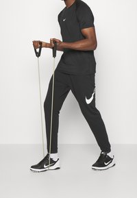 Czarna koszulka sportowa Nike z logo, w połączeniu z czarnymi spodniami z białym logo. Osoba trzyma gumy do ćwiczeń, ubrana w czarno-białe buty.