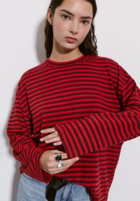 Red and black striped long-sleeve shirt with a crew neckline and oversized sleeves, paired with light blue denim. Silver rings accent the hands.