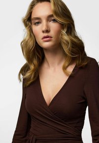 Brun omlottklänning med långa ärmar, djup V-ringning och knytband i midjan. Mjuk textur, figurnära design, minimal detaljering.