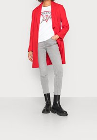 Rød lang frakke over en hvid grafisk t-shirt, parret med lysegrå skinny jeans og sorte kraftige ankelstøvler. Glat stof og skræddersyet design.