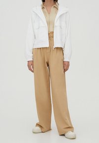 Kobieta ubrana w białą kurtkę z zamkiem, pod którą ma beżowy prążkowany top, luźne brązowe spodnie ze sznurkiem i białe sneakersy na platformie, stojąca na tle jednolitego tła.