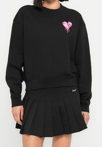 Femme portant un sweat-shirt noir avec un patch cœur rose dégoulinant et une mini-jupe plissée noire sur fond blanc.