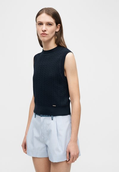 Γυναίκα με ίσια καστανά μαλλιά φοράει αμάνικη μπλούζα πλεκτή σε ναυτικό μπλε και σορτς με κάθετες ρίγες σε ανοιχτό μπλε και άσπρο, σε λευκό φόντο.