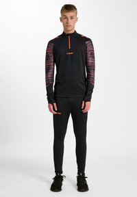 Svart atletisk långärmad topp med röda och grå mönstrade ärmar och halvdragkedja, ihopparad med svarta tajtaleggings och svarta skor.