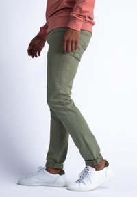 Pantalones verde oliva en un corte ajustado, hechos de una tela suave, con puños enrollados y un bolsillo lateral. Combinados con zapatillas blancas con detalles en verde.