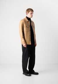 Νέος άνδρας στέκεται με ένα καφετί σακάκι με κουμπιά πάνω από έναν μαύρο ζιβάγκο, μαύρα φαρδιά παντελόνια και μαύρα παπούτσια ενάντια σε έναν απλό λευκό φόντο.