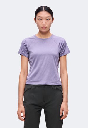 Jeune femme aux cheveux raides et foncés porte un t-shirt lavande à manches courtes et un pantalon noir, debout devant un fond clair uni.