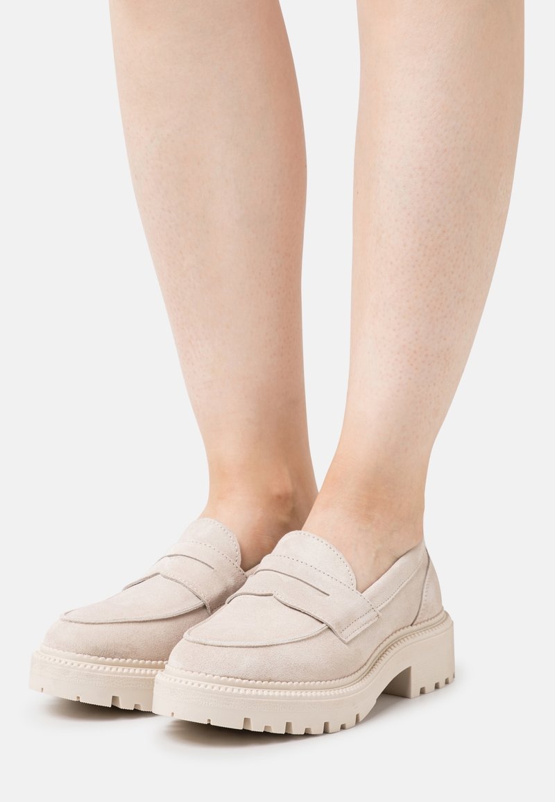 Mocassins épais en daim beige sur une personne à la peau claire, avec des semelles épaisses à crampons et une couture moc toe.