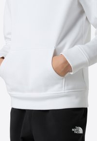 Hanorac alb cu manșete și tiv striate, cu un buzunar frontal tip cangur. Asortat cu pantaloni negri marcați cu un logo în colțul inferior.