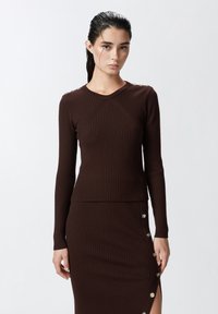 Top lungo a maniche con costine marrone, dalla silhouette aderente, con bottoni decorativi sulla spalla e scollo a girocollo.