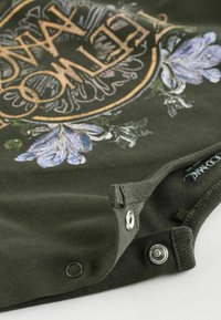 Mørkegrøn bomuldsbeklædning med blomsterdesign i blå, gul cirkel med tekst og sølvknapper i kanten til lukning.