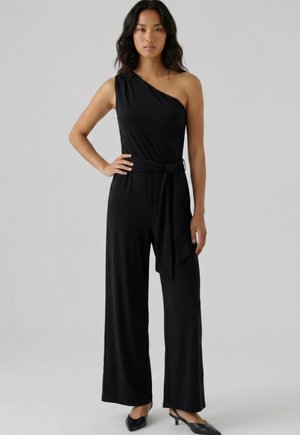 Frau steht mit der Hand auf der Hüfte, trägt einen schwarzen Ein-Schulter-Jumpsuit mit weitem Bein und gebundener Taille, kombiniert mit schwarzen Pumps.