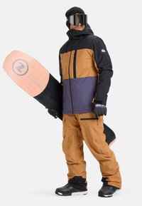 Snowboarder, der eine schwarze, braune und lila Jacke mit Reißverschlüssen, passende braune Hose und schwarze Stiefel trägt, während er ein pinkes und schwarzes Snowboard hält.