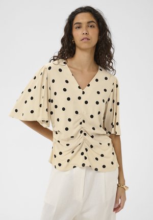 Žena oblečená v béžovej blúzke s výstrihom do V, ktorá má čierne bodky a riasený detail vpredu, v kombinácii s bielymi nohavicami a náramkom.