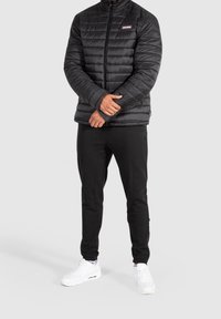 Giacca puffer nera isolata con design a cuciture verticali, colletto alto e zip frontale. Abbinata a pantaloni joggers neri e sneaker bianche.