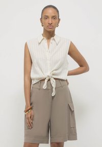 Eine Frau mit goldenen Creolen trägt ein ärmelloses weißes Hemd mit Knöpfen, das an der Taille gebunden ist, und beige Cargo-Shorts, während sie mit einer Hand auf der Hüfte posiert.