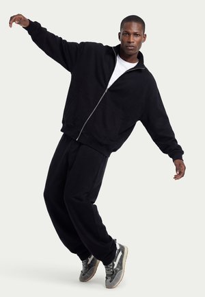 Man wearing black zip-up jacket and pants leans sideways balancing on gray and white sneakers against a plain background.