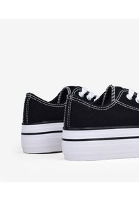 Scarpe da ginnastica nere con lacci bianchi, suola in gomma e cuciture bianche a contrasto. Presentano un design con suola a strati spessa con accenti di strisce nere.