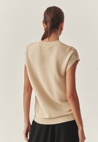 Adīta bēša džemperis ar īsām piedurknēm, ribveida apakšmalu un izšūtām plecu detaļām. Gluda tekstūra, pieguļošs siluets un smalks spīdums.