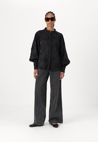 Blusa nera ricamata con maniche a palloncino, abbinata a pantaloni in denim grigi a gamba larga e ballerine nere. Gli accessori includono occhiali da sole oversize.