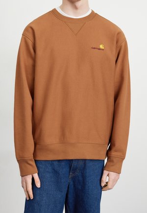 Mand iført en brun Carhartt sweatshirt med ribstrikkede manchetter og kant, kombineret med blå jeans mod en ensfarvet baggrund.
