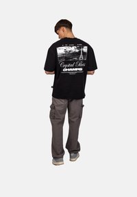 Muž v černém grafickém tričku s potiskem Eiffelovy věže, šedých cargo kalhotách a šedých teniskách, stojící zády k bílé ploše.