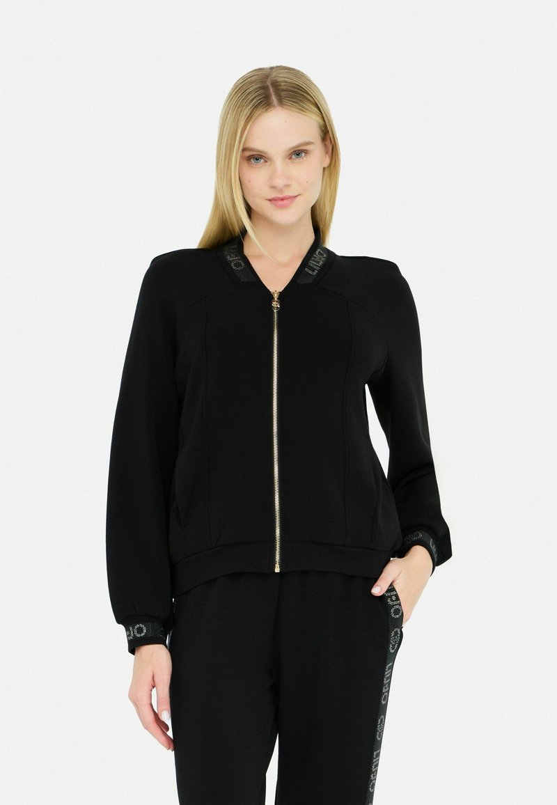 Veste noire zippée avec un col haut, des poignets côtelés et des détails de logo sur le col. Tissu texturé avec une coupe ajustée.
