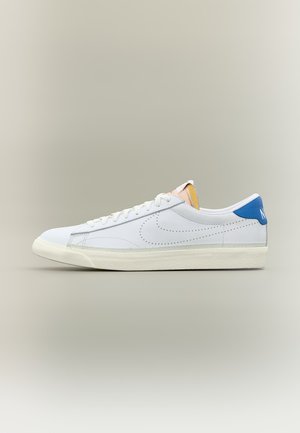 Baskets blanches basses avec un swoosh Nike perforé, un passant de talon bleu, une semelle crème et des lacets blancs sur un fond neutre.