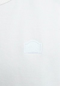Tissu côtelé blanc avec un petit patch rectangulaire blanc brodé présentant un bord supérieur subtilement incurvé centré sur le matériau.