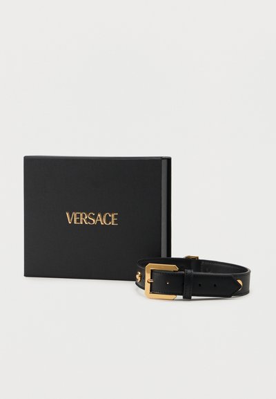 Μαύρη δερμάτινη ζώνη με χρυσή αγκράφα και μαύρο κουτί με την επωνυμία Versace σε χρυσά γράμματα, πάνω σε λευκό φόντο.