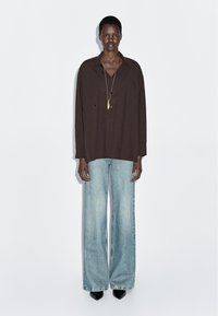 Camicia oversize marrone con bottoni, jeans a gamba larga blu chiaro e scarpe nere a punta. Design semplice con una vestibilità rilassata. Maniche lunghe visibili.