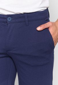 Navy blue trousers with a textured fabric, featuring a button closure, side pockets, and a straight leg design. Subtle branding visible.