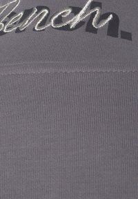 Grauer Strickstoff mit vertikaler Rippung, verziert mit schwarz und metallisch silber gesticktem Text, der "Bench." auf der Oberfläche liest.
