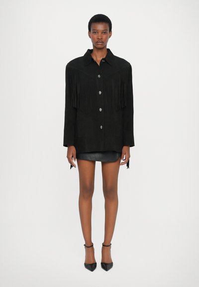 Giacca in suede nera con dettagli in frange sulle spalle, chiusura con bottoni e due tasche anteriori. Abbinata a una gonna in pelle nera aderente.