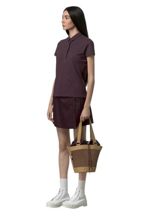 Vrouw met lang donker haar die een paarse polo-jurk met korte mouwen draagt, witte hoge sneakers, en een beige gevlochten handtas met schouderbanden vasthoudt.