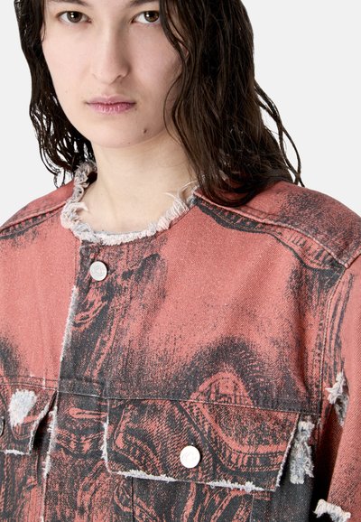 Jeune personne aux cheveux mouillés et foncés portant une veste rose et noire usée avec des bords effilochés et des boutons-pression métalliques.