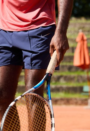 Tennis racket with a blue and black frame, yellow strings, and a textured grip, held by a hand near a pink shirt and navy shorts.