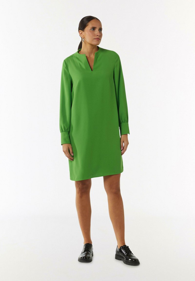 Grünes Kleid aus glattem Stoff, mit V-Ausschnitt und langen Ärmeln mit Manschetten. Kombiniert mit glänzenden schwarzen Schnürschuhen.