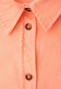 Gros plan sur une chemise en velours côtelé couleur pêche avec un col pointu et trois boutons bruns sur la patte de boutonnage avant.