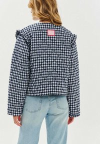 Pikowana kurtka w granatowo-białą krateczkę z falbanami na ramionach, z czerwoną etykietą z tyłu. W połączeniu z jasnoniebieskimi dżinsami.
