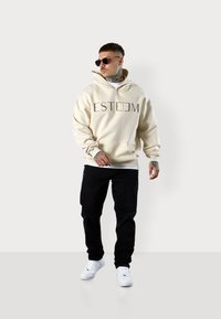 Sweat à capuche crème avec le logo "ESTEEM", associé à un pantalon noir en détresse et des baskets blanches. Modèle portant des lunettes de soleil, tatouages visibles.