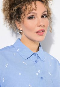 Camicia azzurro chiaro con colletto, decorata con ricami floreali bianchi, presenta una chiusura a bottoni e una tasca a toppa sul lato sinistro.