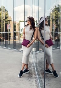 Beige vypasované šaty bez rukávů s bočním rozparkem; drží bordó kabelku přes rameno. Černé tenisky s bílou podrážkou. Reflexní skleněné pozadí.