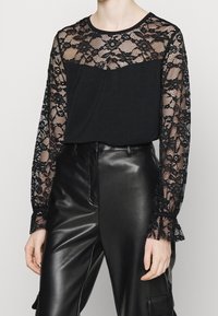 Haut noir à manches longues avec un overlay en dentelle, présentant des motifs floraux, un col rond et un corps en tissu lisse, associé à un pantalon en cuir noir.