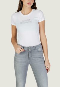 Vrouw die een nauwsluitend wit Armani Exchange T-shirt draagt en lichtgrijze, hooggesneden spijkerbroek, met haar linkerhand die de riemlus van de spijkerbroek vasthoudt.