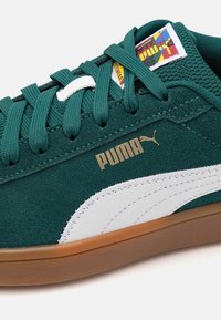 Sneaker verde in suede con dettagli in pelle bianca, logo "PUMA" dorato, lacci rigati e suola in gomma, con un'etichetta colorata sulla linguetta.