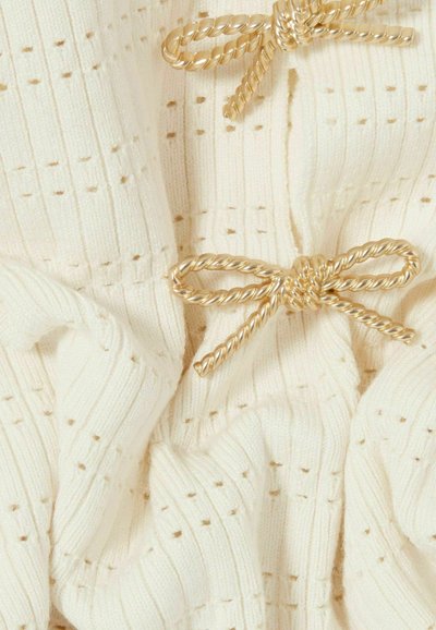 Prenda de punto color crema con textura acanalada y pequeños agujeros uniformemente espaciados. Presenta dos lazos dorados retorcidos como acentos.